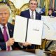 US President Donald Trump holds up a document with his signature in the Oval Office. Begin him is Secretary of the Interior Doug Burghum and a large map showing the US and Canada and the Great Lakes, with Lake Ontario renamed in red script as Lake America.
