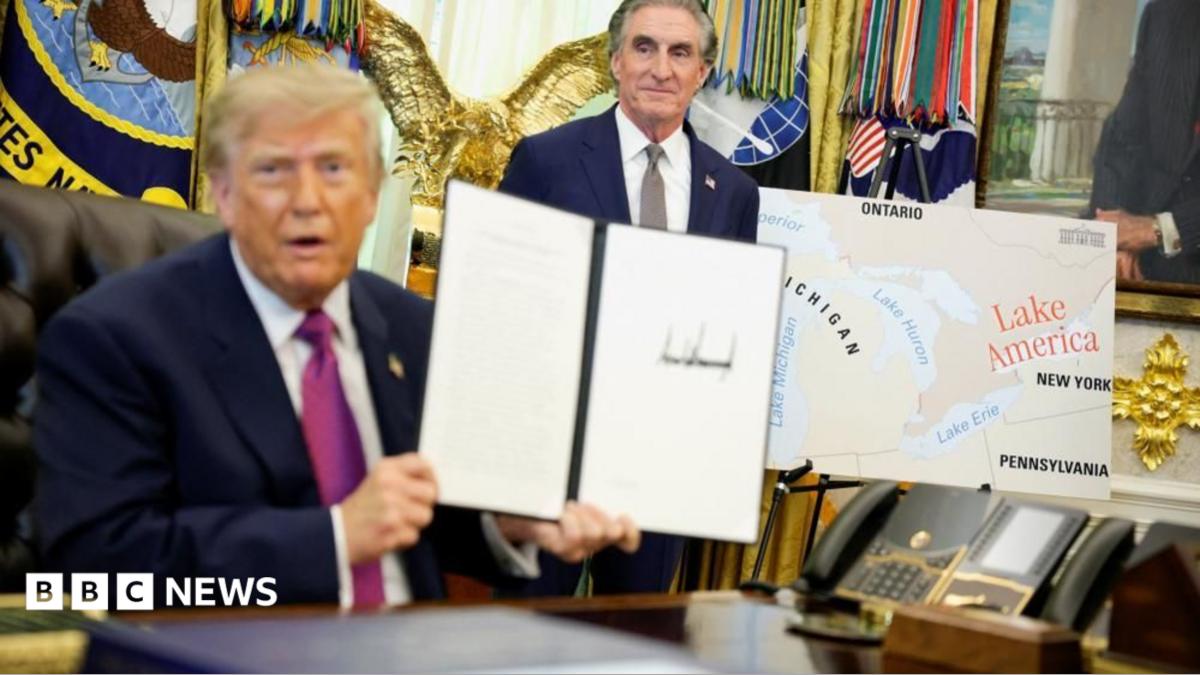 US President Donald Trump holds up a document with his signature in the Oval Office. Begin him is Secretary of the Interior Doug Burghum and a large map showing the US and Canada and the Great Lakes, with Lake Ontario renamed in red script as Lake America.