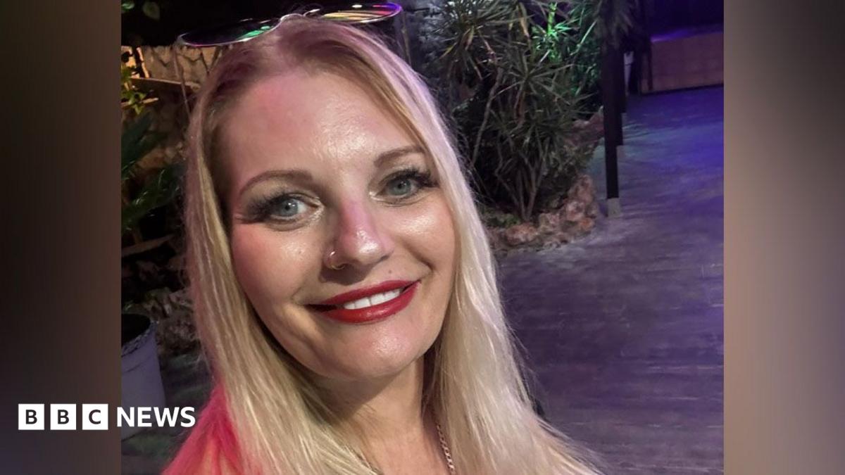 Head-and-shoulders of Michaela Wilson who has long, straight blonde hair, blue eyes, red lipstick and has a pair of sunglasses on top of her head.