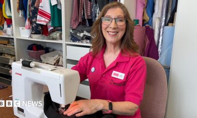 A woman with long brown hair and glasses is sitting at a sewing machine and smiling at the camera. She is wearing a pink collared shirt with the Dressability logo on it