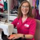 A woman with long brown hair and glasses is sitting at a sewing machine and smiling at the camera. She is wearing a pink collared shirt with the Dressability logo on it