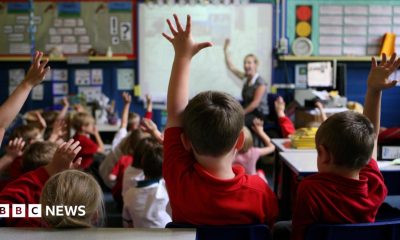 Children sit in a primary school classroom with several pupils raising their hands while a teacher stands at the front near an interactive whiteboard. School displays and learning materials can be seen around the room.