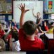Children sit in a primary school classroom with several pupils raising their hands while a teacher stands at the front near an interactive whiteboard. School displays and learning materials can be seen around the room.