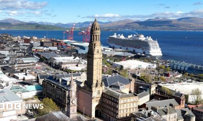 A coastal cityscape with a tall clock tower in the foreground, surrounded by historic and modern buildings. Behind it, a large white cruise ship is docked beside red industrial cranes. Beyond the harbour, green hills and mountains rise under a partly cloudy blue sky.