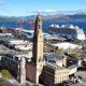 A coastal cityscape with a tall clock tower in the foreground, surrounded by historic and modern buildings. Behind it, a large white cruise ship is docked beside red industrial cranes. Beyond the harbour, green hills and mountains rise under a partly cloudy blue sky.