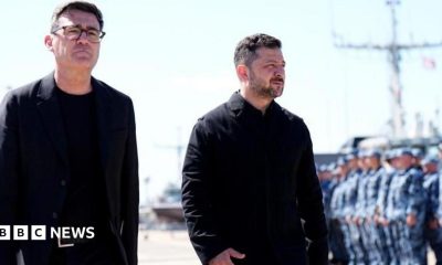 Andy Burnham and Volodymyr Zelensky walk next to each other at a naval base. Both men are dressed in black and have short, dark hair. Burnham is wearing glasses. Military personnel can be seen in the background.