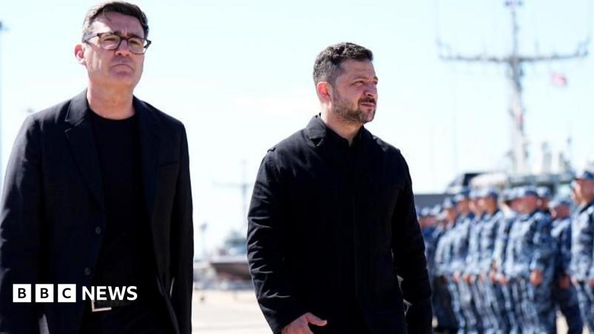 Andy Burnham and Volodymyr Zelensky walk next to each other at a naval base. Both men are dressed in black and have short, dark hair. Burnham is wearing glasses. Military personnel can be seen in the background.