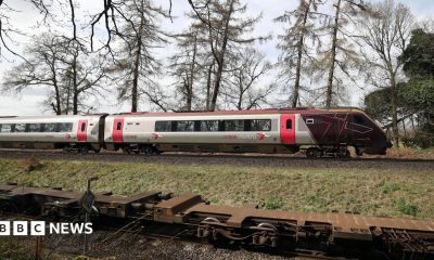 The cab and first carriage of a CrossCountry train travels on tracks in the countryside. In the background trees and fields can be seen.