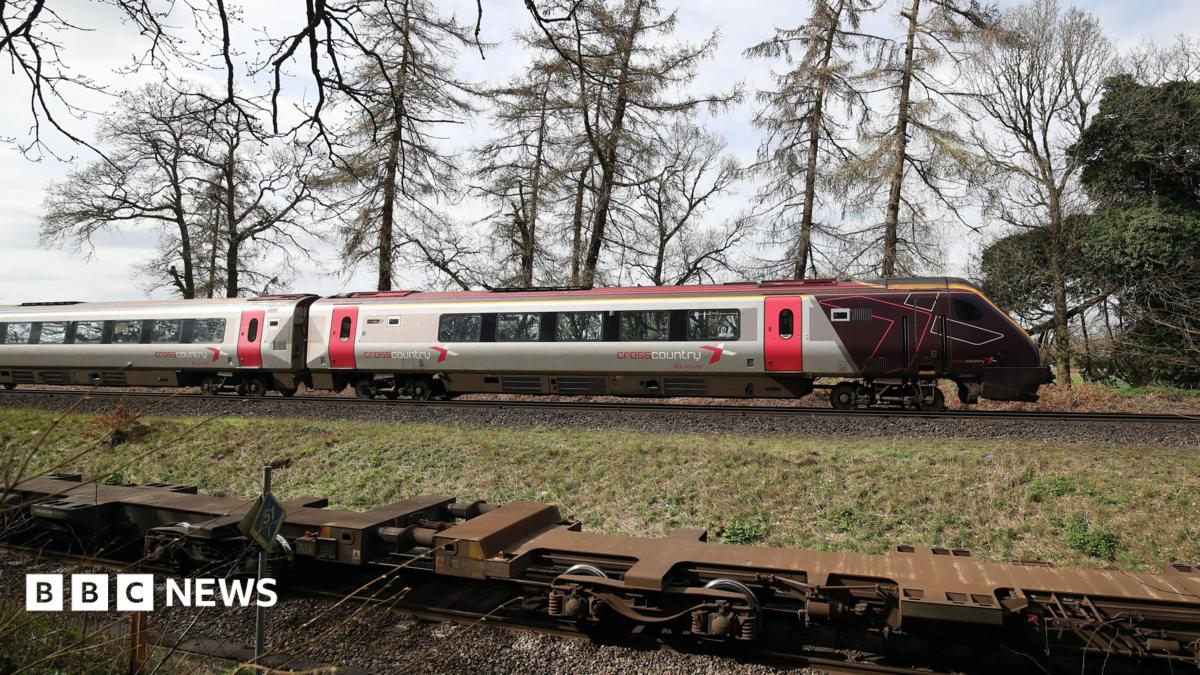 The cab and first carriage of a CrossCountry train travels on tracks in the countryside. In the background trees and fields can be seen.