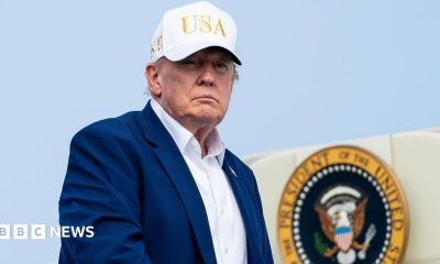 Trump, wearing a suit and no tie and a white "USA" hat, descending from airplane steps