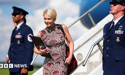 Natalie Harp walks off Air Force One wearing a sleeveless printed red dress. She is smiling and has a laptop and several phones in her hands.