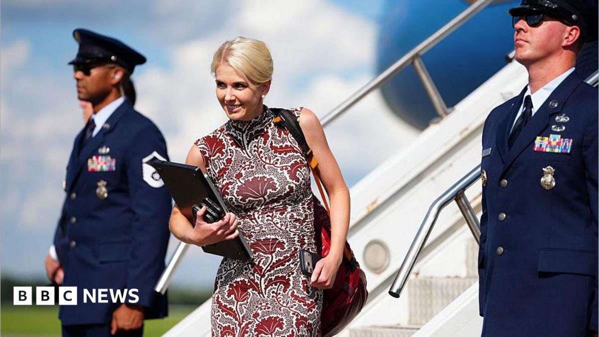 Natalie Harp walks off Air Force One wearing a sleeveless printed red dress. She is smiling and has a laptop and several phones in her hands.