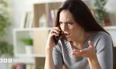 Woman with angry expression holds phone up to her ear looking frustrated. She has brown hair and wears a grey knit top.