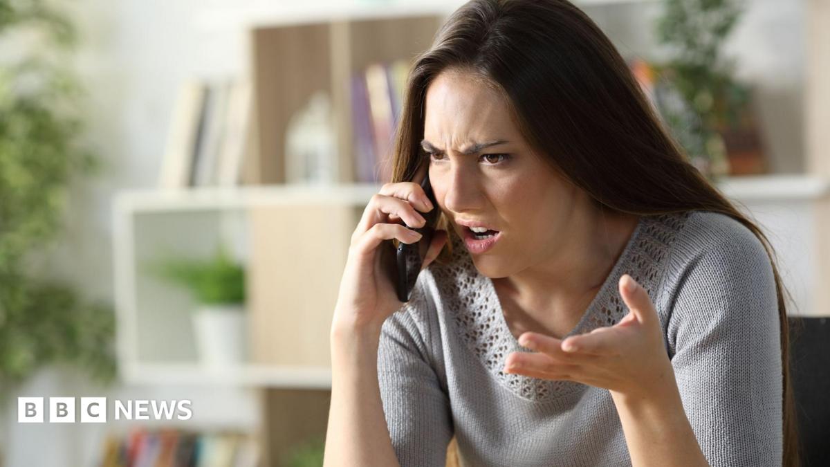 Woman with angry expression holds phone up to her ear looking frustrated. She has brown hair and wears a grey knit top.