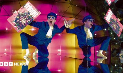 Finatan McCarney poses during the launch of the 2026 Edinburgh Festival Fringe programme at Camera Obscura. He is wearing a blue suit, white scarf and sunglasses and his reflection is repeated across the image.