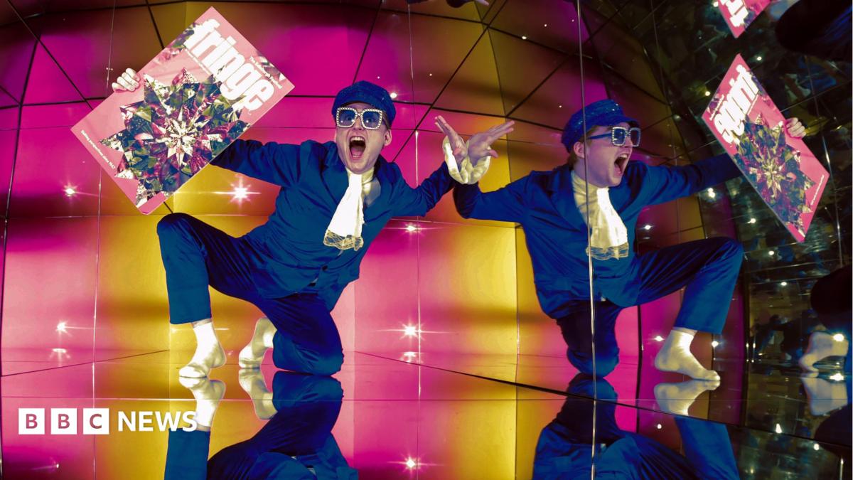 Finatan McCarney poses during the launch of the 2026 Edinburgh Festival Fringe programme at Camera Obscura. He is wearing a blue suit, white scarf and sunglasses and his reflection is repeated across the image.