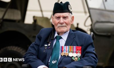 Donald Turrell is sitting in a wheelchair in front of a military vehicle. He is wearing a black army hat and a suit. On the left side of his jacket is a row of medals from World War Two.