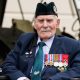 Donald Turrell is sitting in a wheelchair in front of a military vehicle. He is wearing a black army hat and a suit. On the left side of his jacket is a row of medals from World War Two.