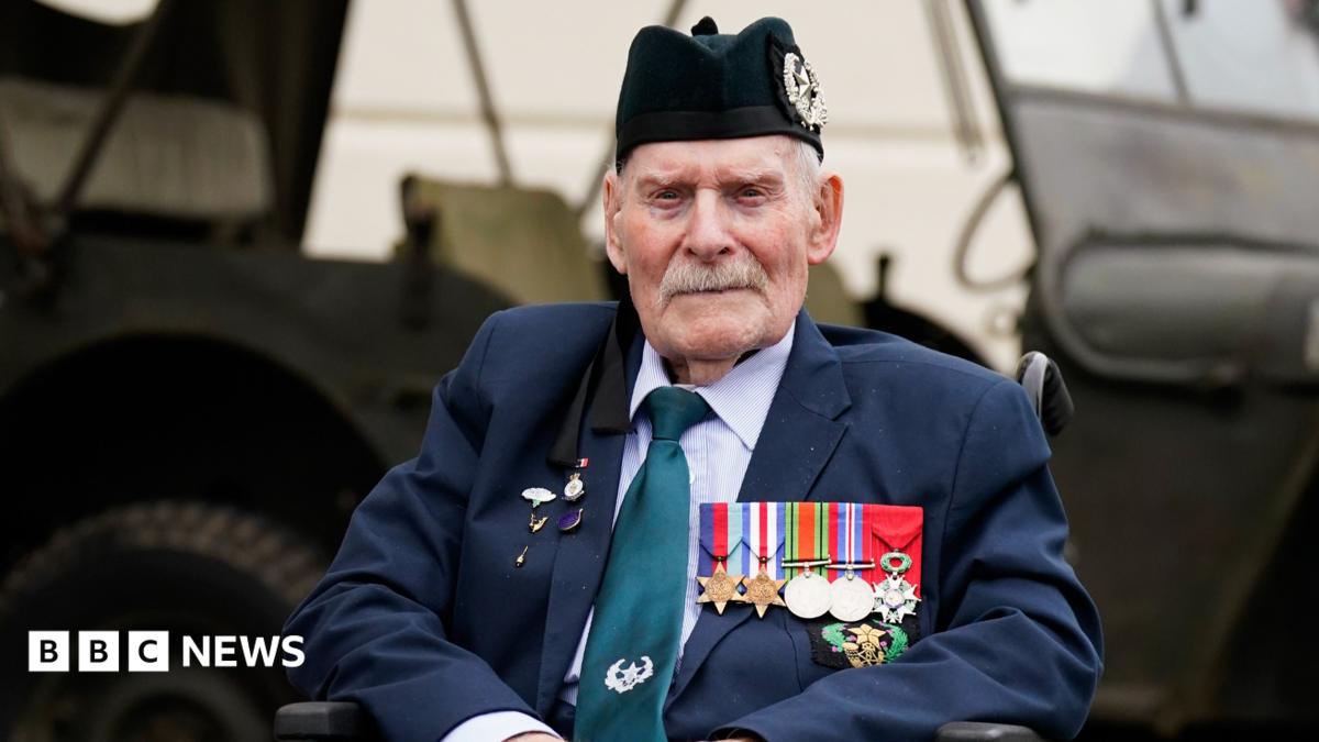 Donald Turrell is sitting in a wheelchair in front of a military vehicle. He is wearing a black army hat and a suit. On the left side of his jacket is a row of medals from World War Two.