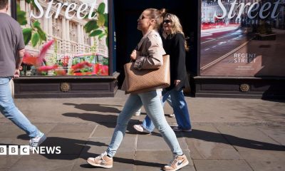 A woman in jeans and carrying a large tote bag walking along a high street. She is seen in profile against the backdrop of a shop window in London and other shoppers are in the background
