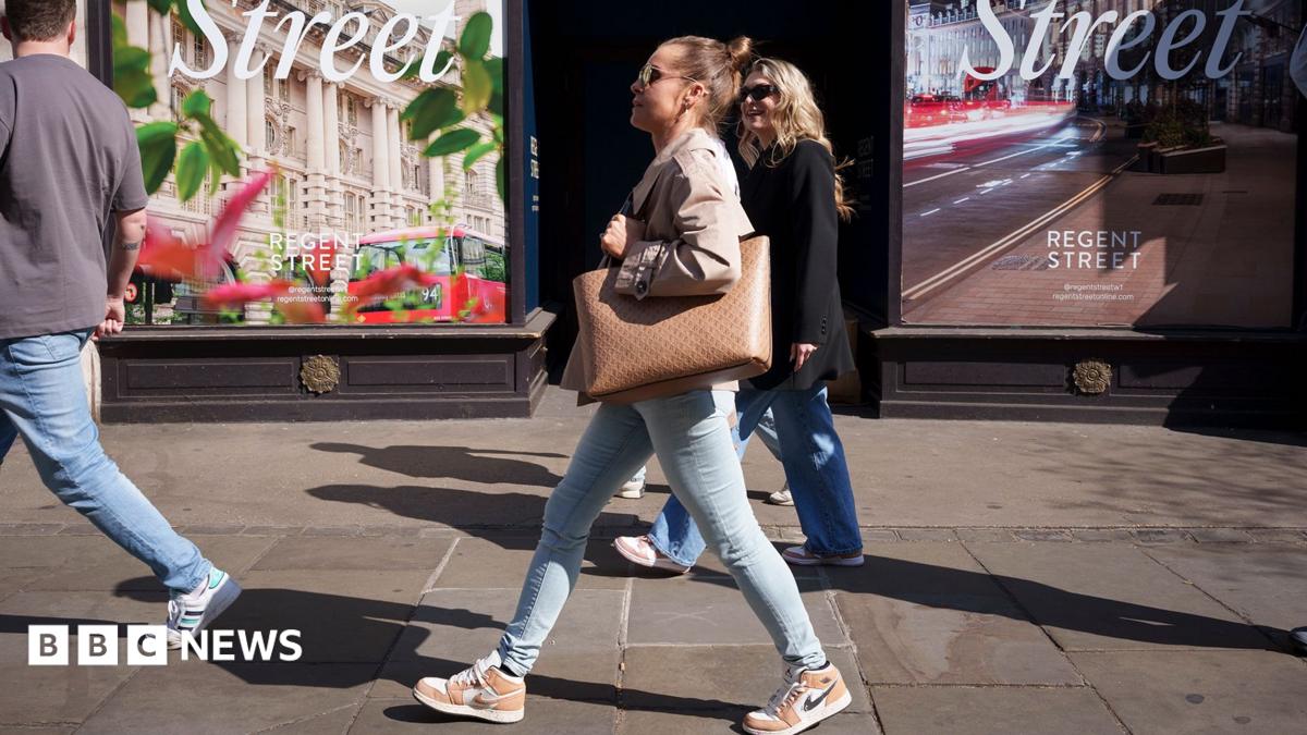 A woman in jeans and carrying a large tote bag walking along a high street. She is seen in profile against the backdrop of a shop window in London and other shoppers are in the background
