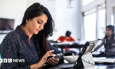 A female office worker sitting in front of a laptop and looking at her phone