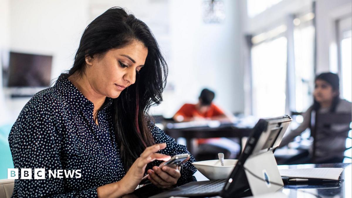 A female office worker sitting in front of a laptop and looking at her phone