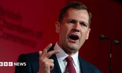 Robert Jenrick makes a hand expression as he speaks at a podium in front of a red-lit background.