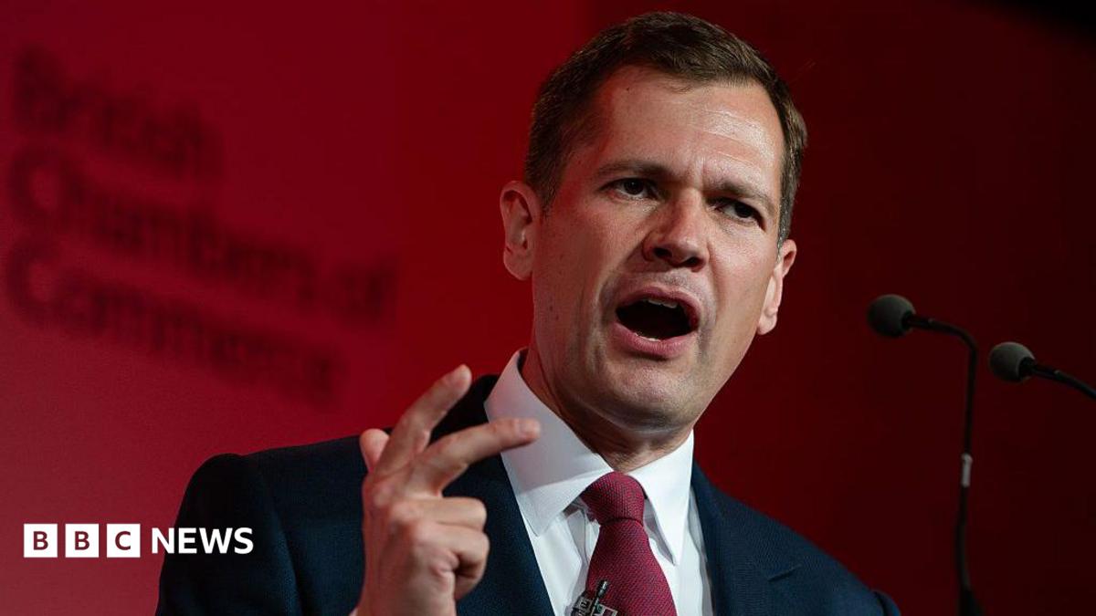Robert Jenrick makes a hand expression as he speaks at a podium in front of a red-lit background.