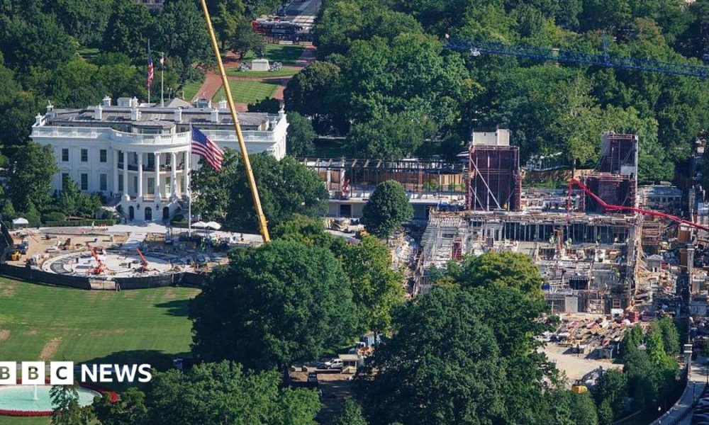An aerial image of the White House, a large white building with tall classic columns. To the right of the building there's a large construction site, with scaffolding and cranes building a new structure.