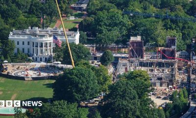 An aerial image of the White House, a large white building with tall classic columns. To the right of the building there's a large construction site, with scaffolding and cranes building a new structure.