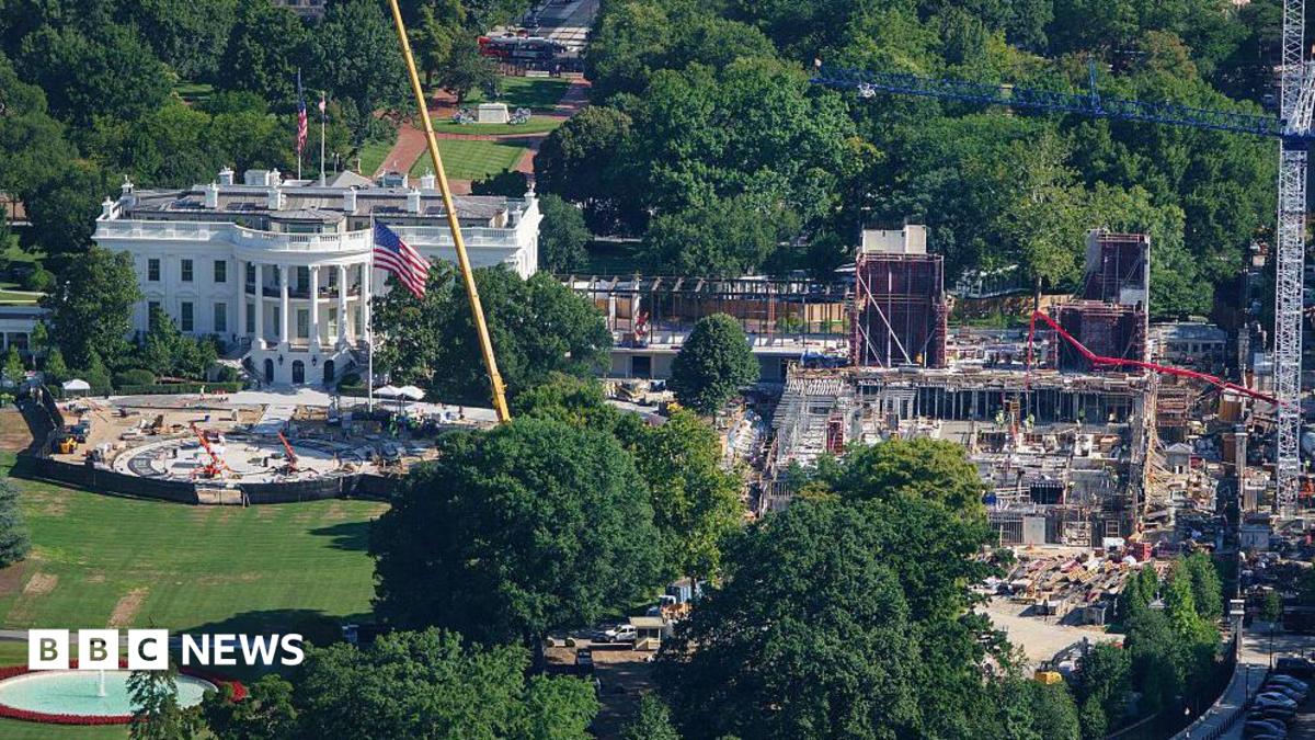 An aerial image of the White House, a large white building with tall classic columns. To the right of the building there's a large construction site, with scaffolding and cranes building a new structure.