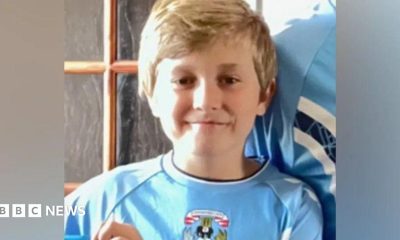 A boy in a sky blue Coventry City home shirt is smiling at the camera