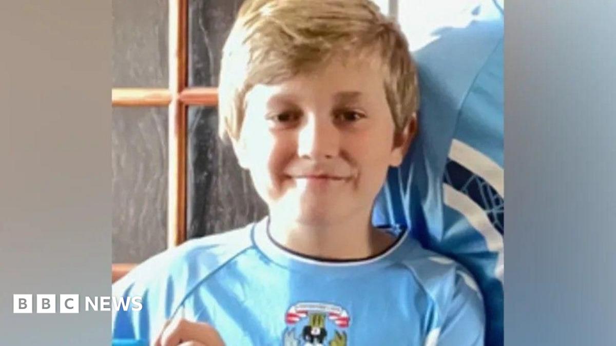 A boy in a sky blue Coventry City home shirt is smiling at the camera