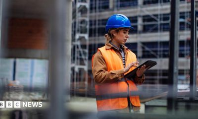 A young woman working on a building site. She is wearing a blue hard hat and an orange hi-viz jacket over her clothes. She is looking at an iPad.