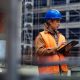 A young woman working on a building site. She is wearing a blue hard hat and an orange hi-viz jacket over her clothes. She is looking at an iPad.