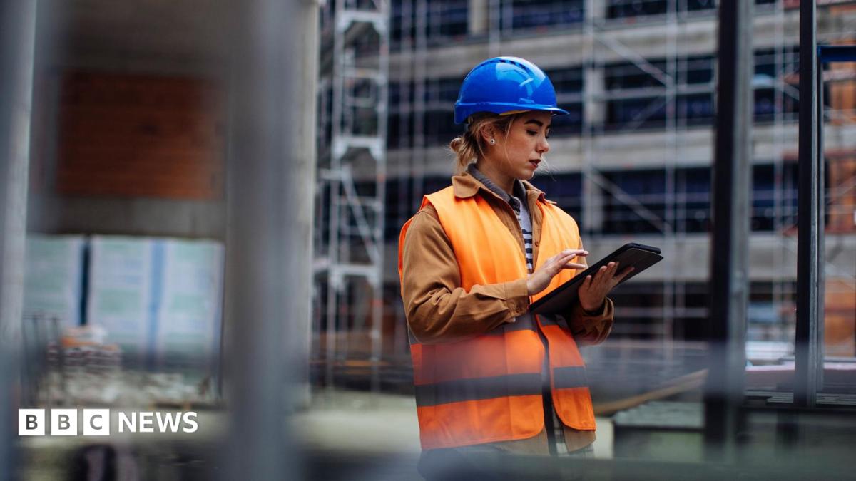 A young woman working on a building site. She is wearing a blue hard hat and an orange hi-viz jacket over her clothes. She is looking at an iPad.