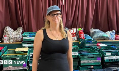 Zia Lee stands in a black vest top with a denim cap on and black glasses and long blonde hair.  Behind her are green crates with school names and carefully folded uniforms in them.