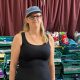 Zia Lee stands in a black vest top with a denim cap on and black glasses and long blonde hair.  Behind her are green crates with school names and carefully folded uniforms in them.