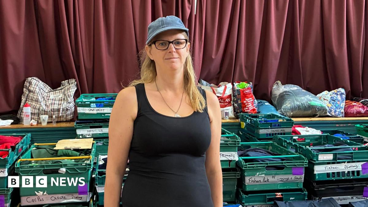 Zia Lee stands in a black vest top with a denim cap on and black glasses and long blonde hair.  Behind her are green crates with school names and carefully folded uniforms in them.