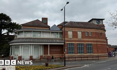 A large building on the corner of a town street with large windows that are covered with grey paint. Part of the building is red brick and has blacked out windows with blur frames. There is a small tower with a circular logo with the letters "BHS" on it