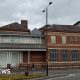 A large building on the corner of a town street with large windows that are covered with grey paint. Part of the building is red brick and has blacked out windows with blur frames. There is a small tower with a circular logo with the letters "BHS" on it