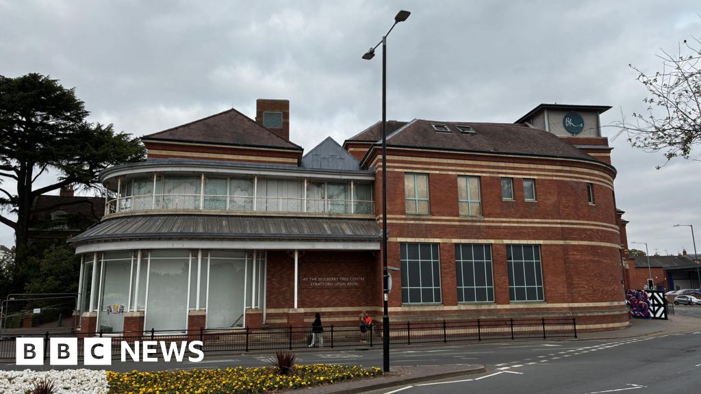 A large building on the corner of a town street with large windows that are covered with grey paint. Part of the building is red brick and has blacked out windows with blur frames. There is a small tower with a circular logo with the letters "BHS" on it