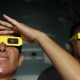 Two people wearing yellow eclipse-viewing glasses in the foreground look upwards. One person raises a hand above the glasses to shade their eyes. The second person is positioned behind them, also looking towards the sky. The scene appears to be indoors, with a dark, blurred background and sunlight illuminating the faces and glasses.