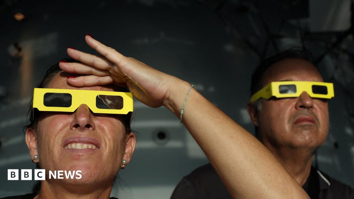 Two people wearing yellow eclipse-viewing glasses in the foreground look upwards. One person raises a hand above the glasses to shade their eyes. The second person is positioned behind them, also looking towards the sky. The scene appears to be indoors, with a dark, blurred background and sunlight illuminating the faces and glasses.