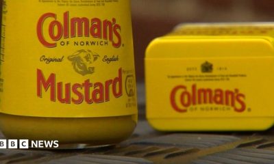 A jar and tin of Colman's Mustard propped up on a surface next to each other. The tin of mustard has been laid on its side. The packaging on each item is a mustard yellow and the label reads, Colman's of Norwich, original English Mustard.