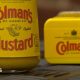A jar and tin of Colman's Mustard propped up on a surface next to each other. The tin of mustard has been laid on its side. The packaging on each item is a mustard yellow and the label reads, Colman's of Norwich, original English Mustard.