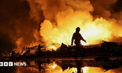 Firefighters work at a site of a Russian missile strike, their silhouettes contrasted against the glowing yellow and orange of smoke plumes illuminated by large fires.