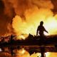 Firefighters work at a site of a Russian missile strike, their silhouettes contrasted against the glowing yellow and orange of smoke plumes illuminated by large fires.
