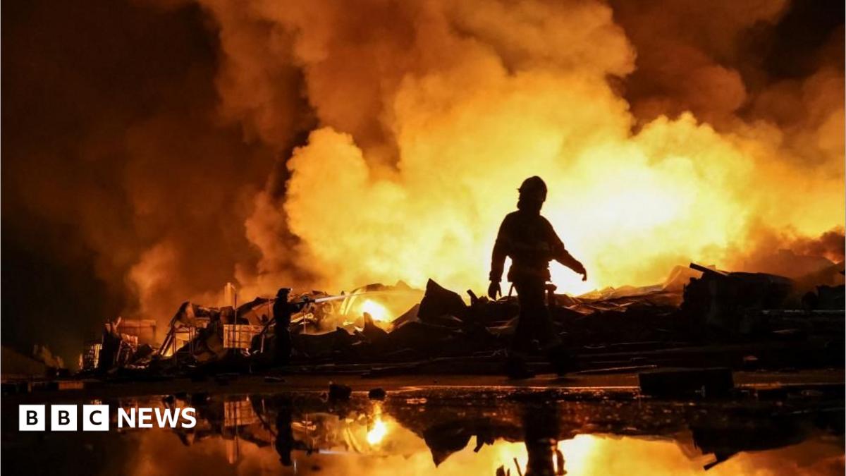 Firefighters work at a site of a Russian missile strike, their silhouettes contrasted against the glowing yellow and orange of smoke plumes illuminated by large fires.